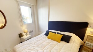 Een keurig opgemaakt bed met een boek erop, een nachtkastje met een lamp, en een raam met jaloezieën.