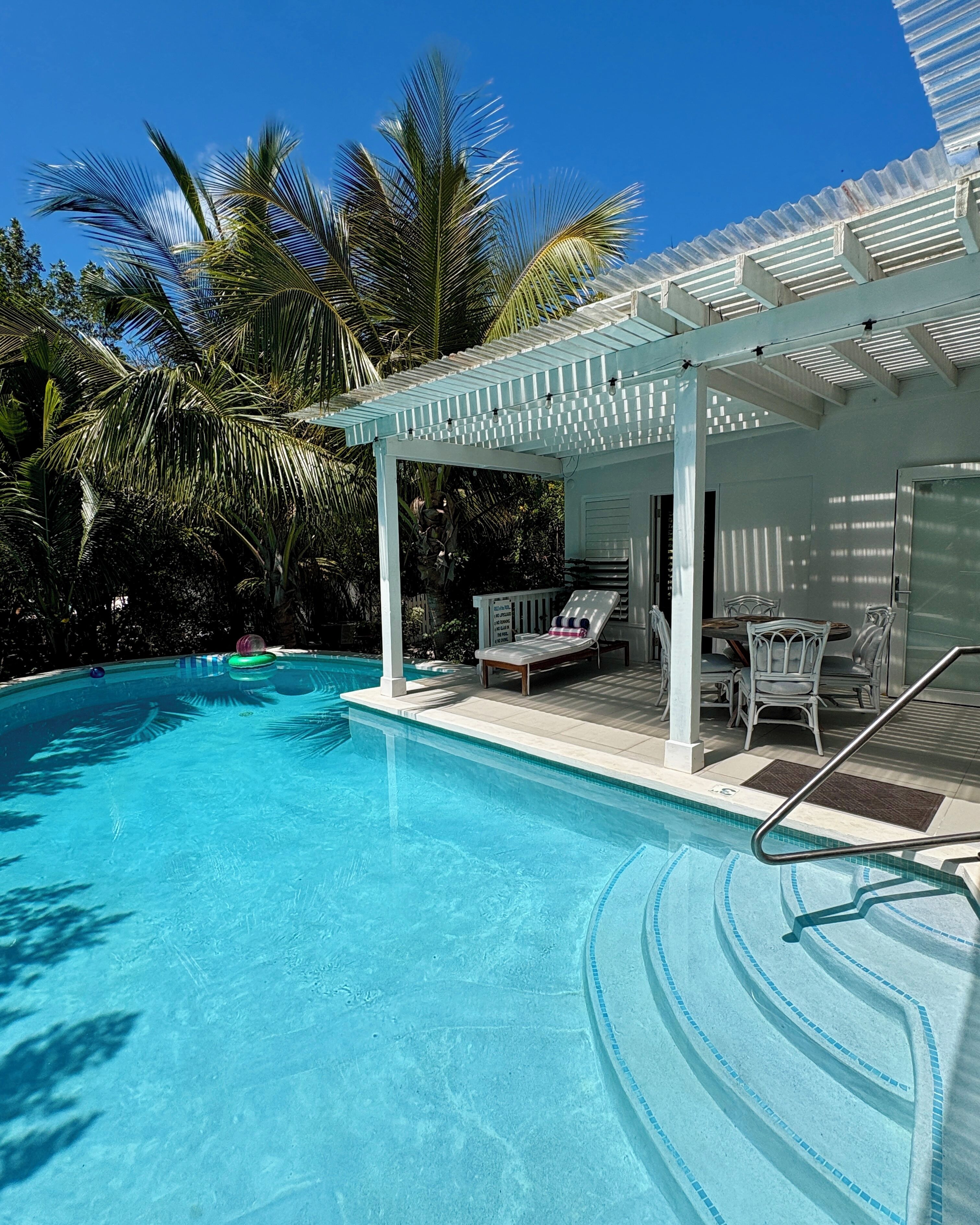 A clear blue pool with a white pergola, lounge chairs, and a small building with a door.