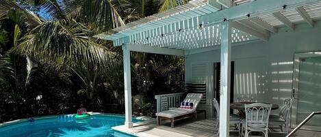 A clear blue pool with a white pergola, lounge chairs, and a small building with a door.