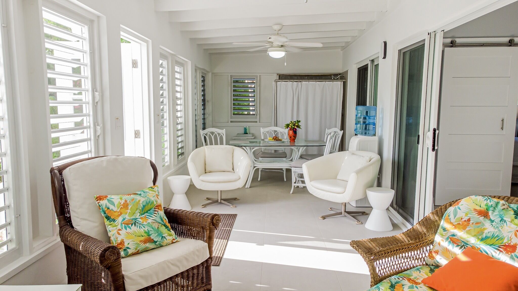 A bright, sunlit room with wicker furniture, a dining table, and a ceiling fan.