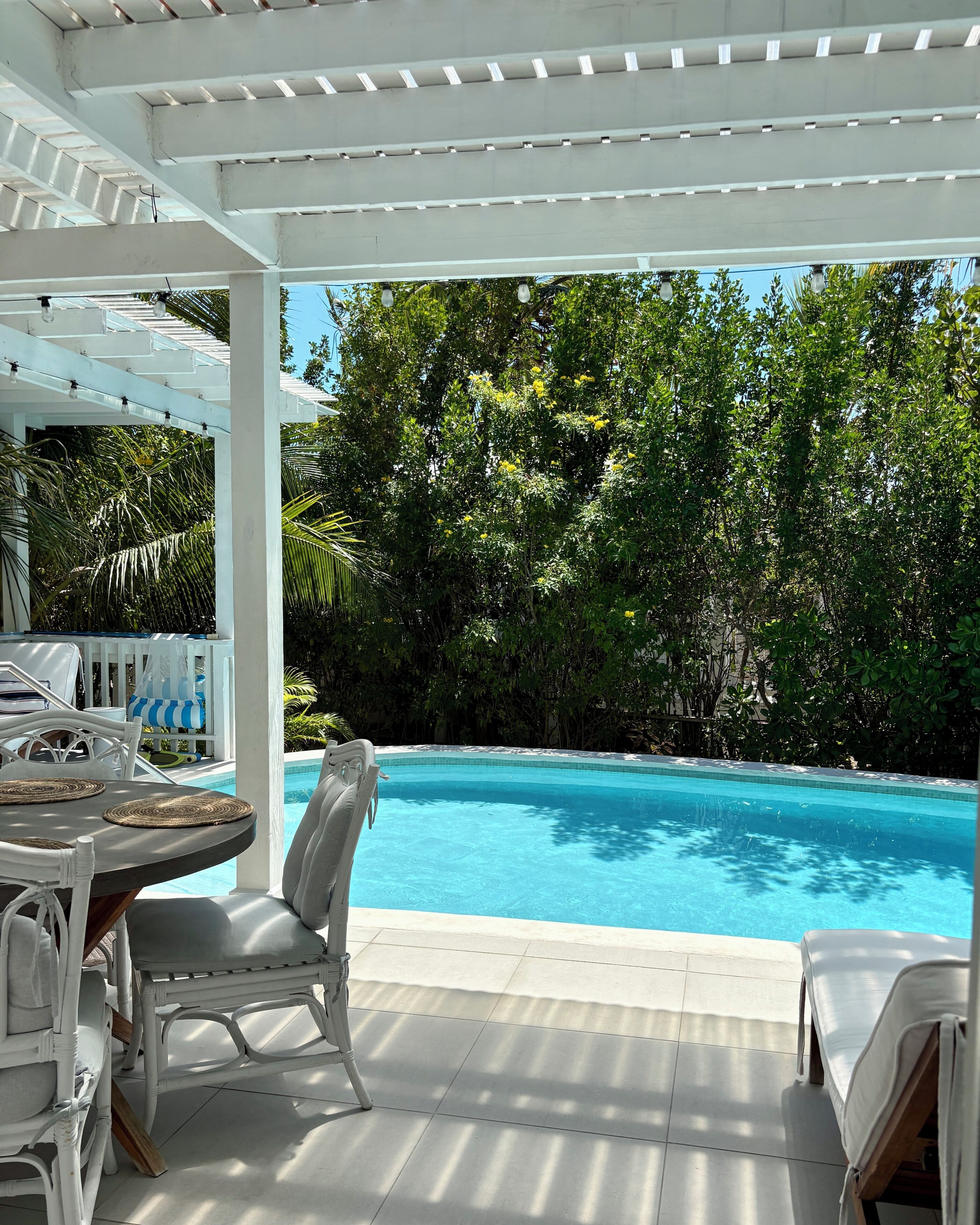A covered outdoor area with a pool, white furniture, and lush greenery.