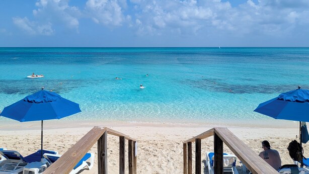 A beach with clear turquoise water, a sandy shore, and wooden deck chairs under blue umbrellas.