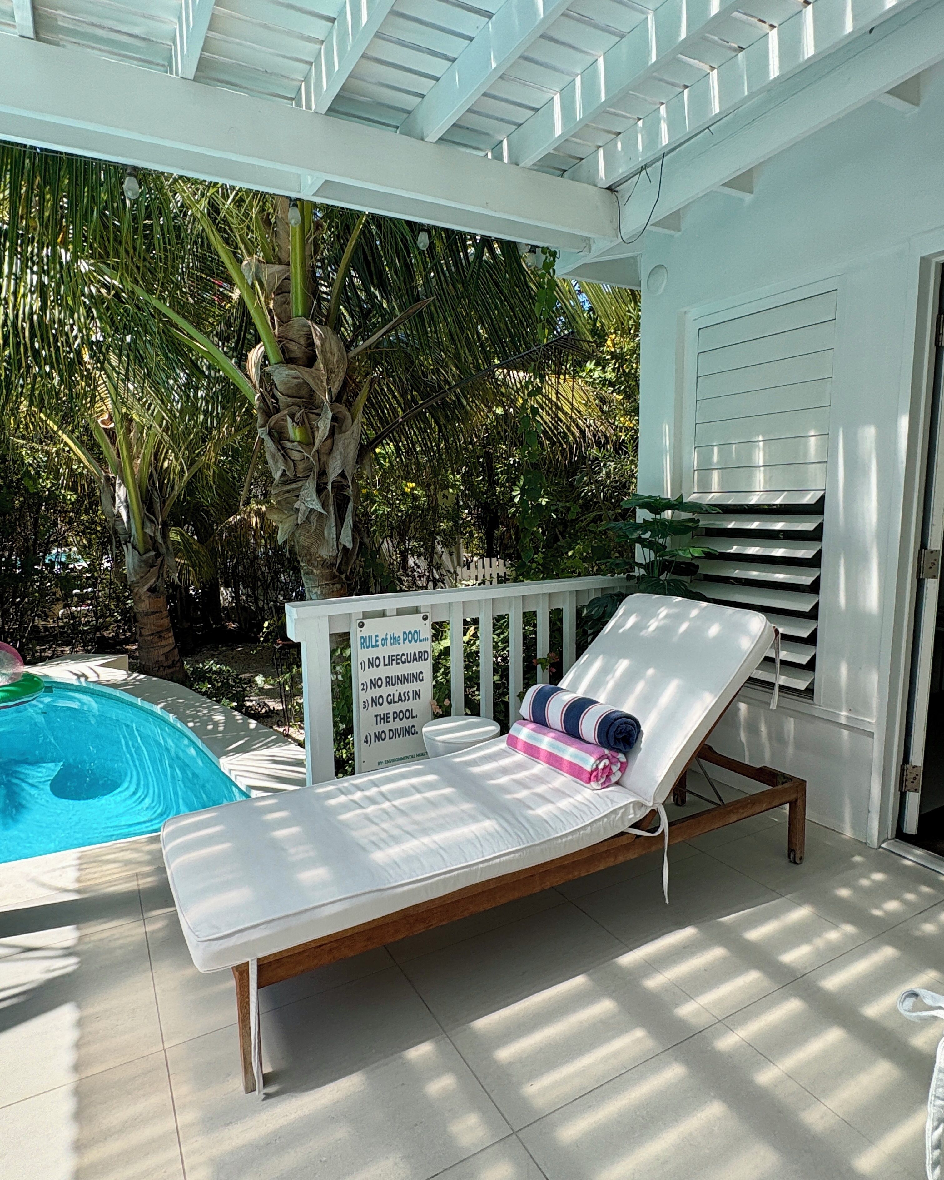 A poolside lounge chair with a white cushion and a towel, a pool with clear blue water, and a sign listing pool rules.