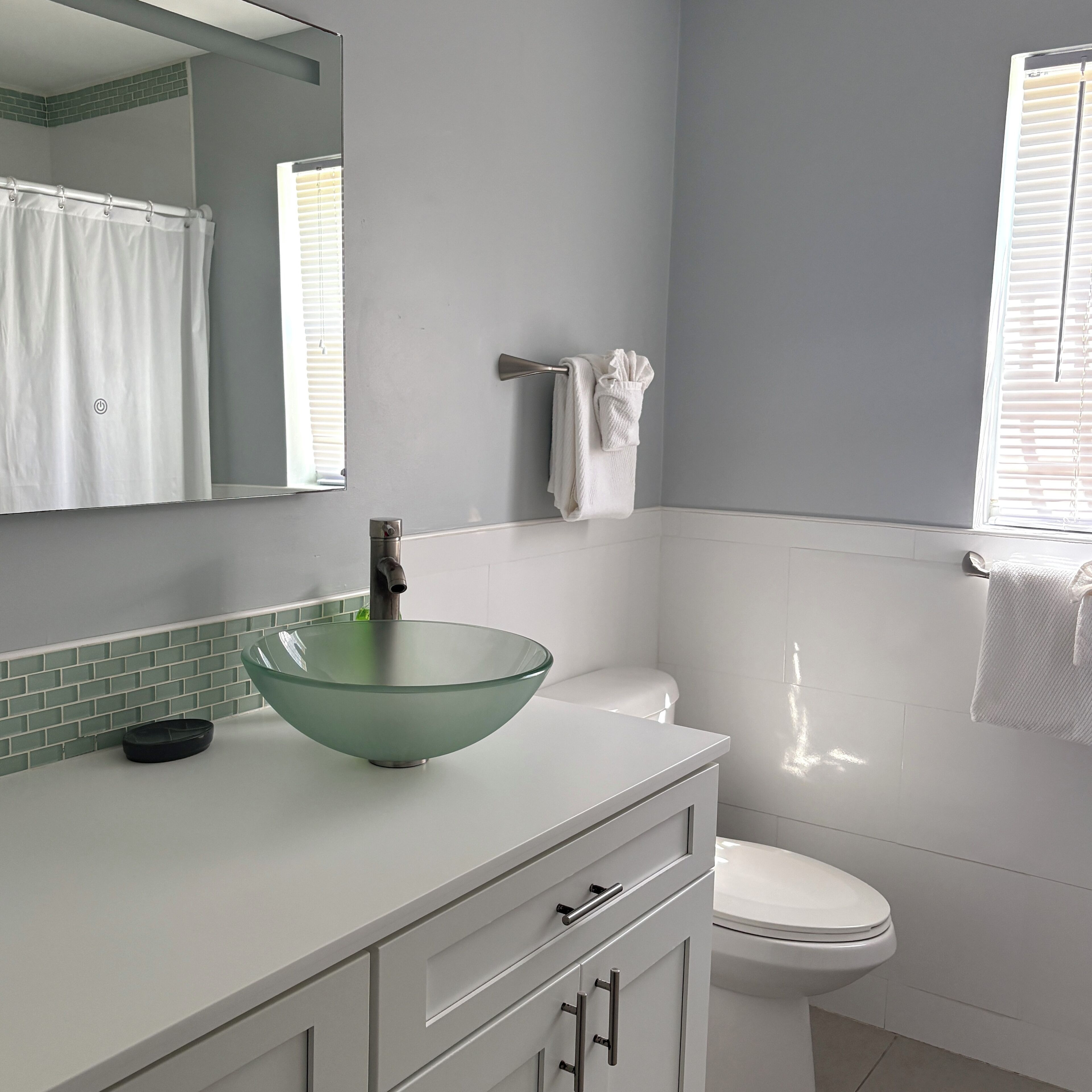 A bathroom with a glass bowl sink, a white toilet, a towel rack, and a window with blinds.