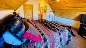 A cozy room with a sloped ceiling, a bed with patterned bedding, a television, and a door. - Relaxing 5 Bedroom Log Cabin with Large Backyard (Donnelly)