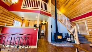 A log cabin interior with a central staircase, wooden walls, a kitchen area, and a fireplace. - Relaxing 5 Bedroom Log Cabin with Large Backyard (Donnelly)