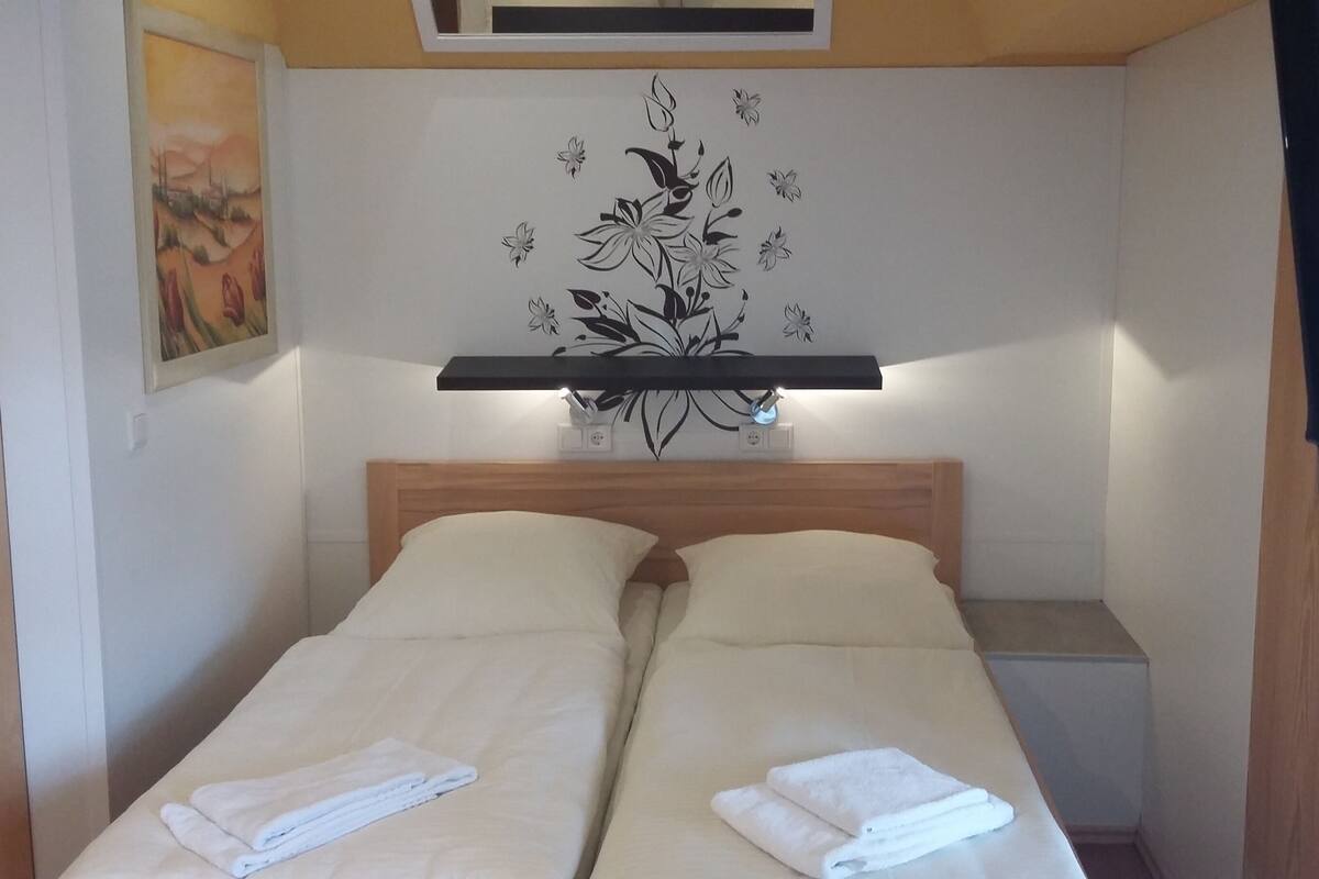Ein Doppelbett mit zwei Kissen und zwei gefalteten Handtüchern, ein an der Wand befestigtes Regal mit floralem Dekor sowie ein Bild an der Wand.