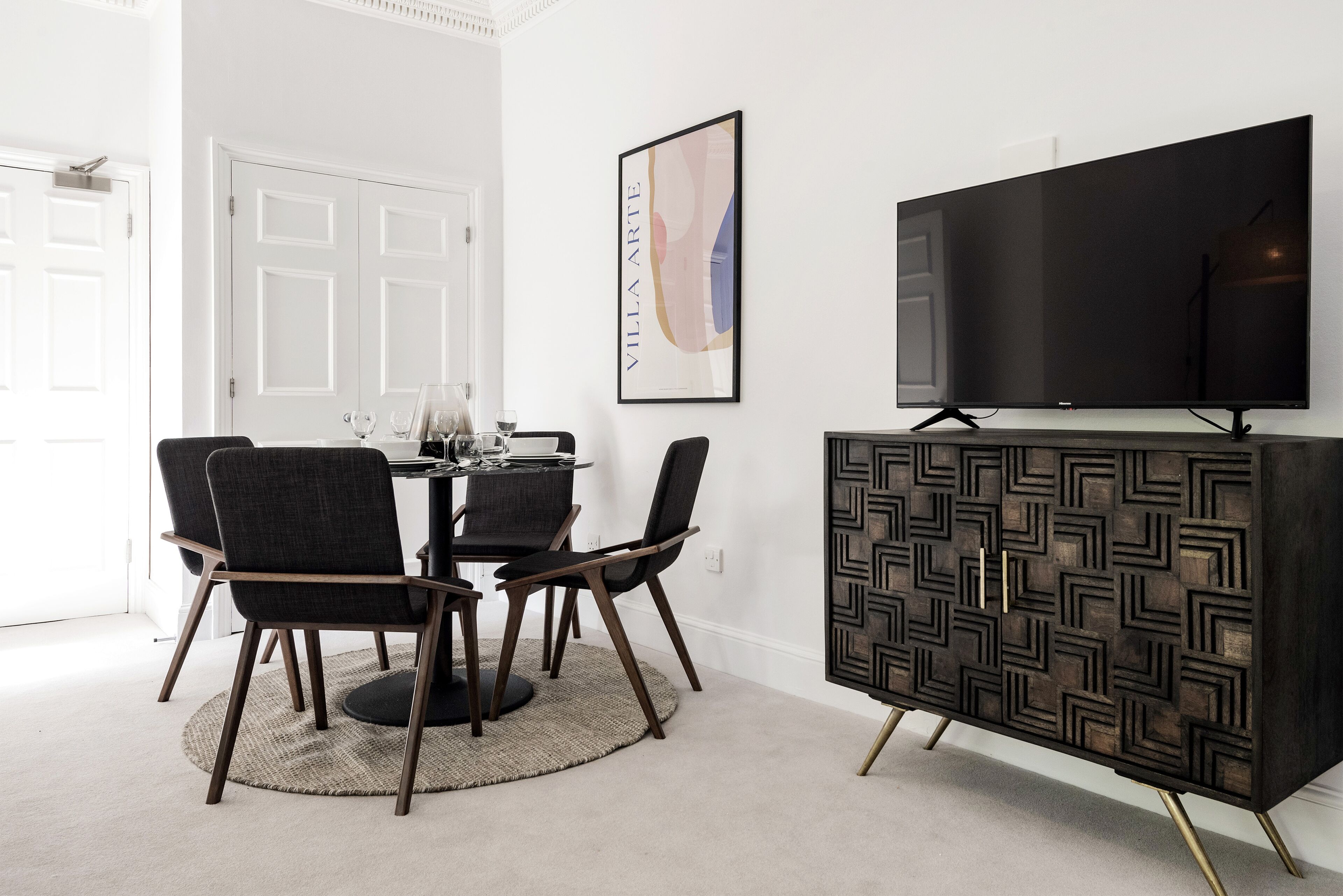 A dining area with a round table, four chairs, a black cabinet, and a framed artwork on the wall.