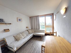 A cozy room with a wooden table, a sofa with patterned cushions, a window with curtains, and a wall-mounted light fixture.