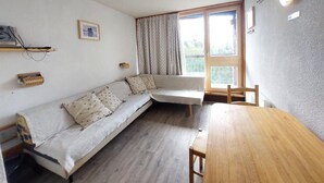 A cozy room with a wooden table, a sofa with patterned cushions, a window with curtains, and a wall-mounted light fixture.