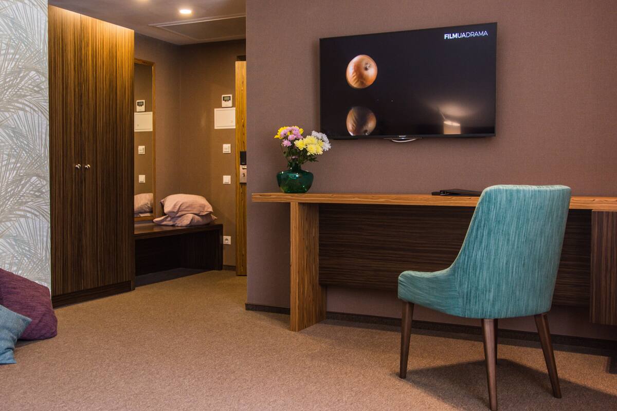 A hotel room with a wooden desk, a teal chair, a flat-screen TV, a green vase with flowers, a wooden wardrobe, and a bed with pink bedding.