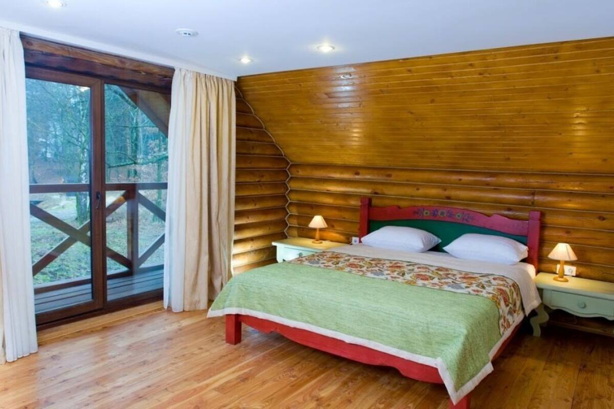 A bedroom with a wooden sloped ceiling, a large window with curtains, a bed with a floral bedspread, and two bedside tables with lamps.