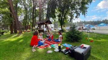 A group of people sitting on a blanket in a park with trees and a gazebo in the background.