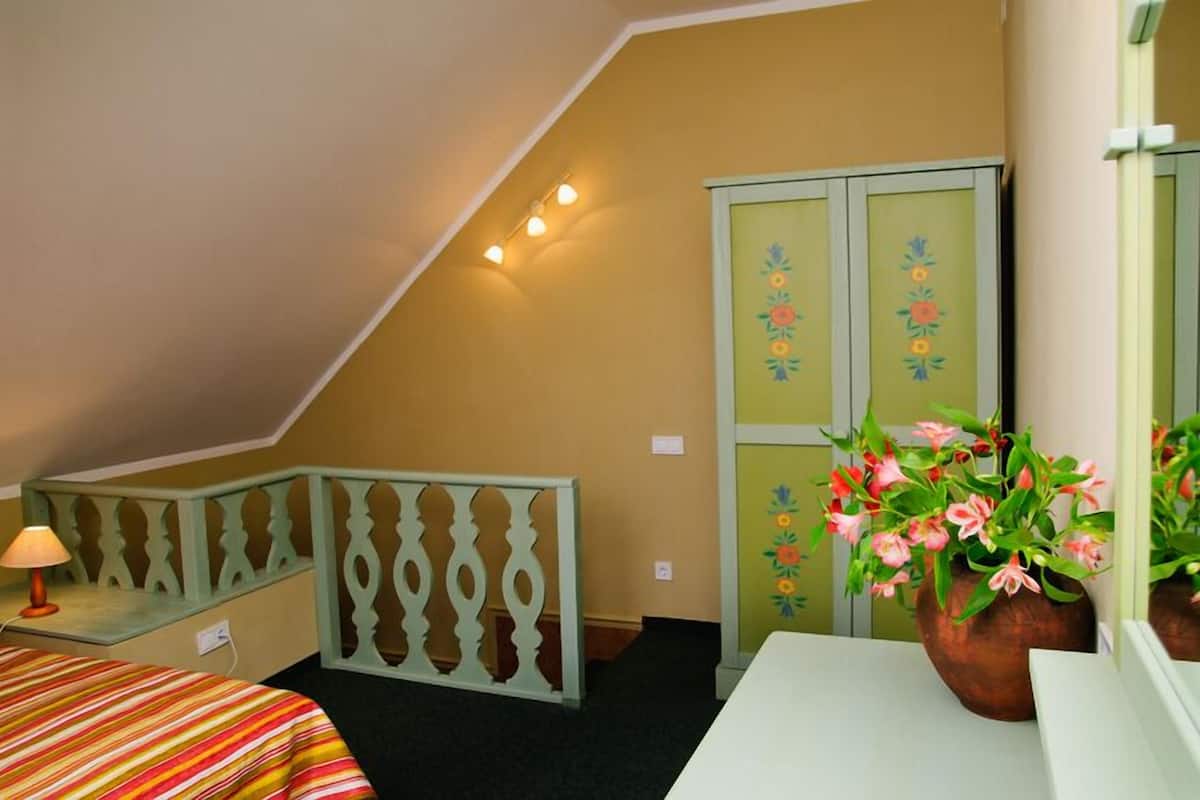 A bedroom with a sloped ceiling, a bed with a striped bedspread, a nightstand with a lamp, a dresser with floral decorations, and a potted plant.