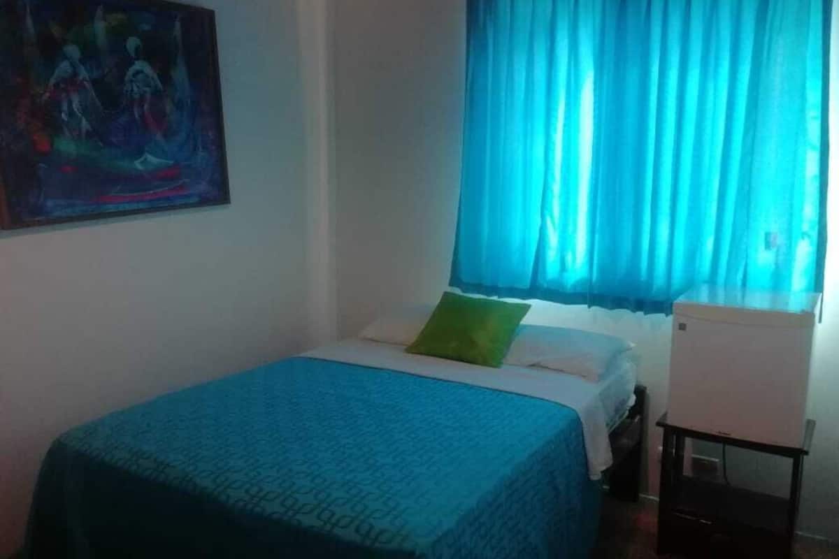A room with a bed, a green pillow, a white bedside table, and a painting on the wall.