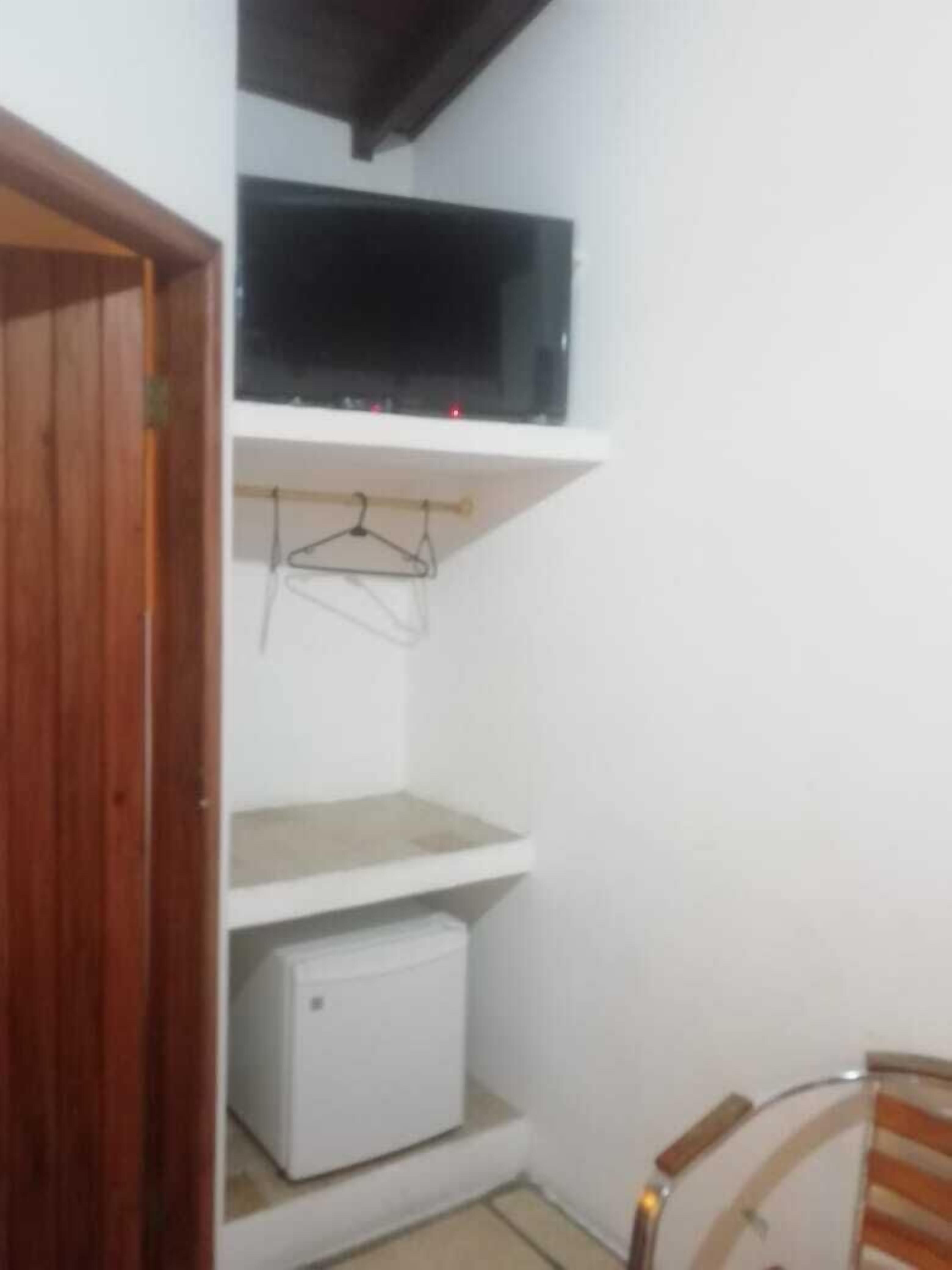 A small room with a wooden door, a white wall, a white shelf with a TV and a white appliance below it, and a chair with a striped cushion.
