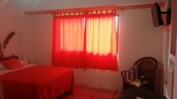 A room with two beds, a chair, a table, and a window with red curtains.