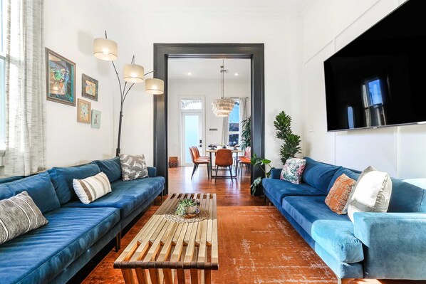 A modern living room with two blue sofas, a wooden coffee table, and a dining area in the background.
