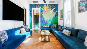 Ein modernes Wohnzimmer mit einem großen Flachbildfernseher, blauen Sofas, einem Holztisch und einem farbenfrohen Wandbild.