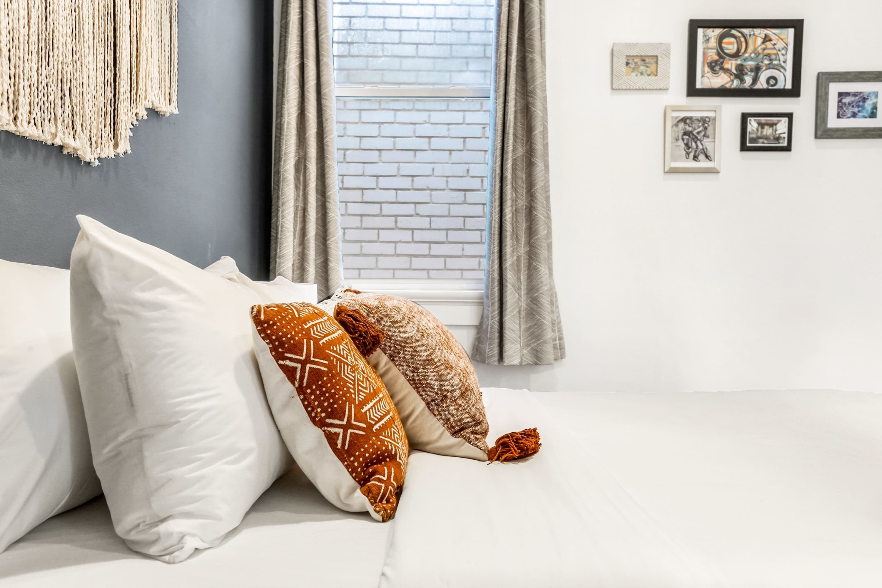 A bed with white and patterned pillows, a macrame wall hanging, and framed artwork on a white wall.