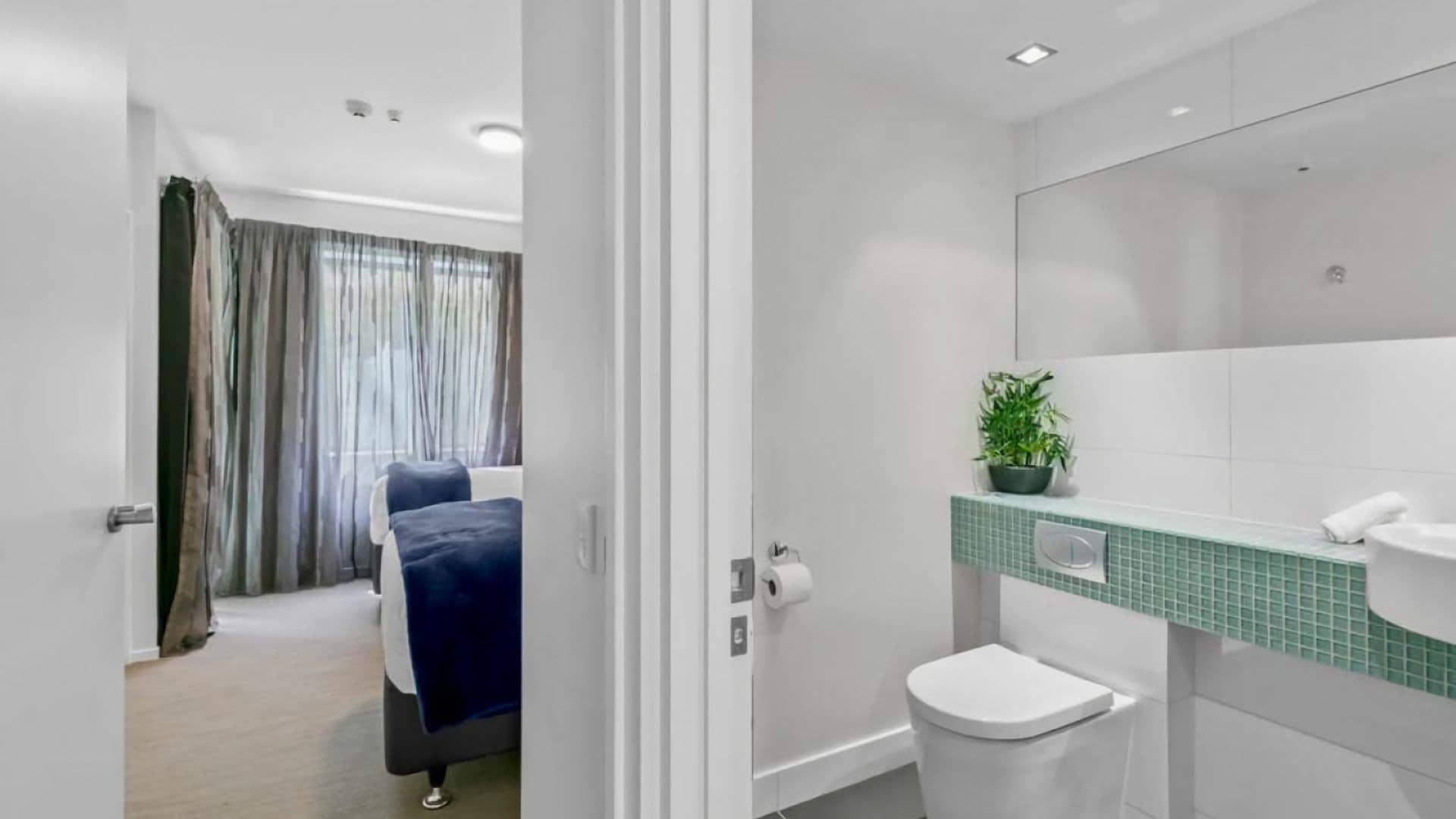 Ein modernes Badezimmer mit Toilette, Waschbecken und Spiegel. Auf der Ablagefläche des Waschbeckens steht eine Pflanze und im Hintergrund ist eine Duschkabine zu sehen.