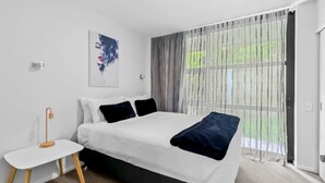 Een moderne slaapkamer met een groot bed, een nachtkastje, een schilderij aan de muur en een uitzicht door dunne gordijnen.
