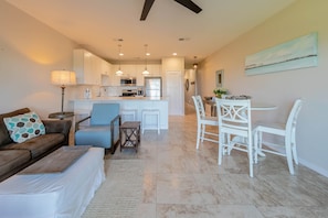 A modern living room with a brown leather sofa, a white ottoman, and a blue armchair. There is a dining area with a white table and chairs, and a kitchen with white cabinets and stainless steel appliances.