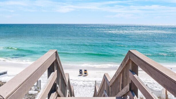 Une vue sur la plage, avec une promenade en bois menant au rivage, un poste de surveillance des sauveteurs et un ciel bleu dégagé.