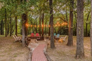 Un espace extérieur aménagé avec des chaises et un foyer, entouré d’arbres et illuminé de guirlandes lumineuses.