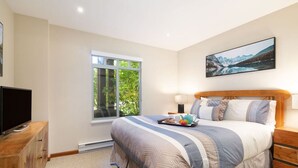 Ein Schlafzimmer mit einem großen Bett, einem Fernseher auf einer Holzkommode, einem Fenster mit Blick ins Grüne und einem Gemälde an der Wand.