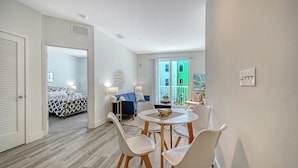 Une chambre d’hôtel moderne avec un lit, un canapé bleu, une table à manger avec des chaises, et une vue sur un bâtiment verdoyant à l’extérieur.