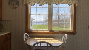 A dining area with a table and chairs, a window with a view of the outdoors, and a clock on the wall. - Oceanfront mid-coast Maine Getaway! (Stockton Springs)