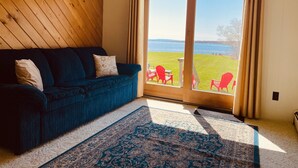 A cozy room with a dark blue sofa, a patterned rug, and a view of a grassy area with red chairs outside. - Oceanfront mid-coast Maine Getaway! (Stockton Springs)