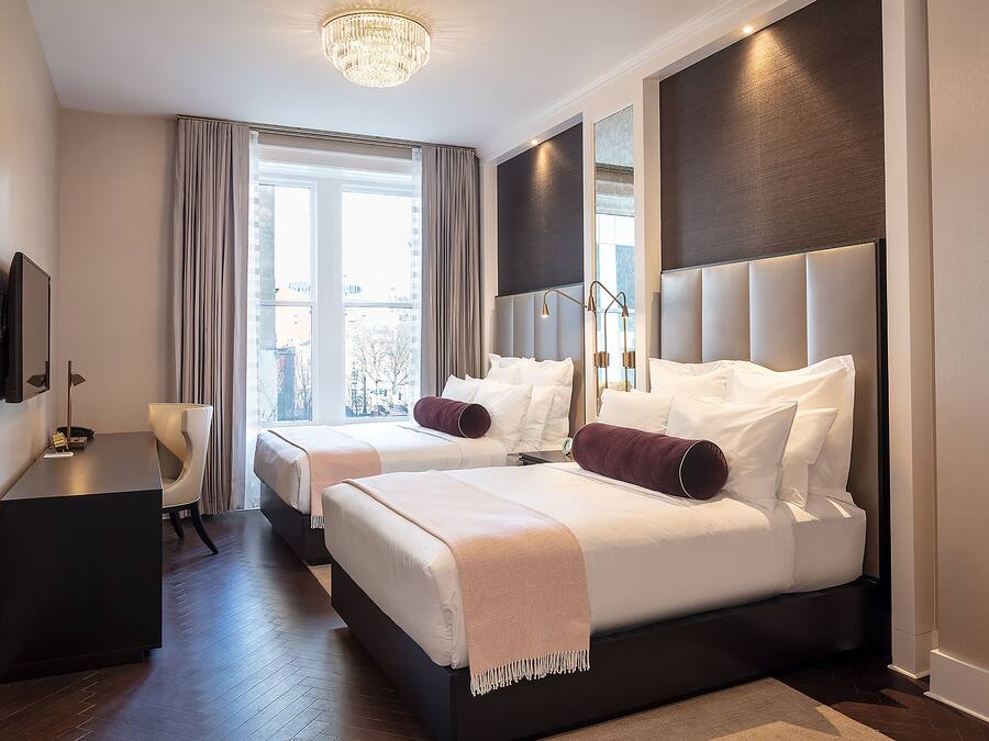 A hotel room with two beds, a desk, a TV, and a large window with curtains.