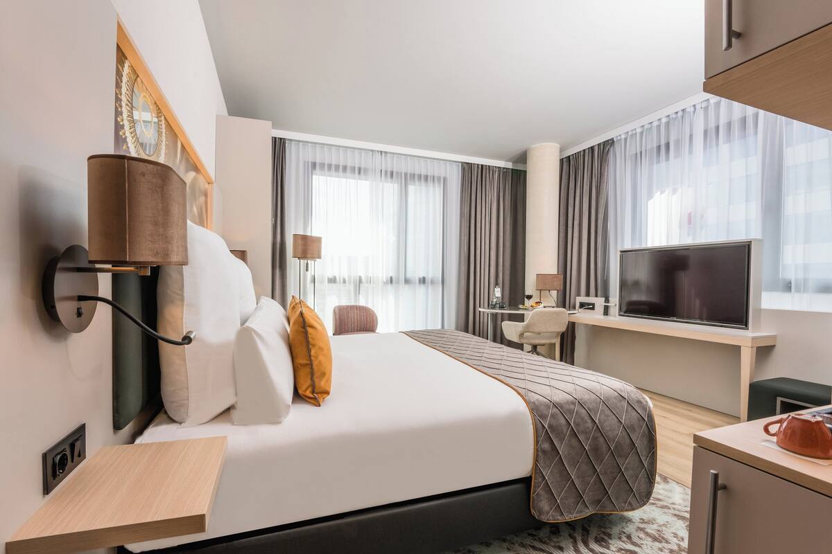 Une chambre d’hôtel moderne équipée d’un lit, d’un bureau, d’une télévision et d’une fenêtre avec des rideaux.
