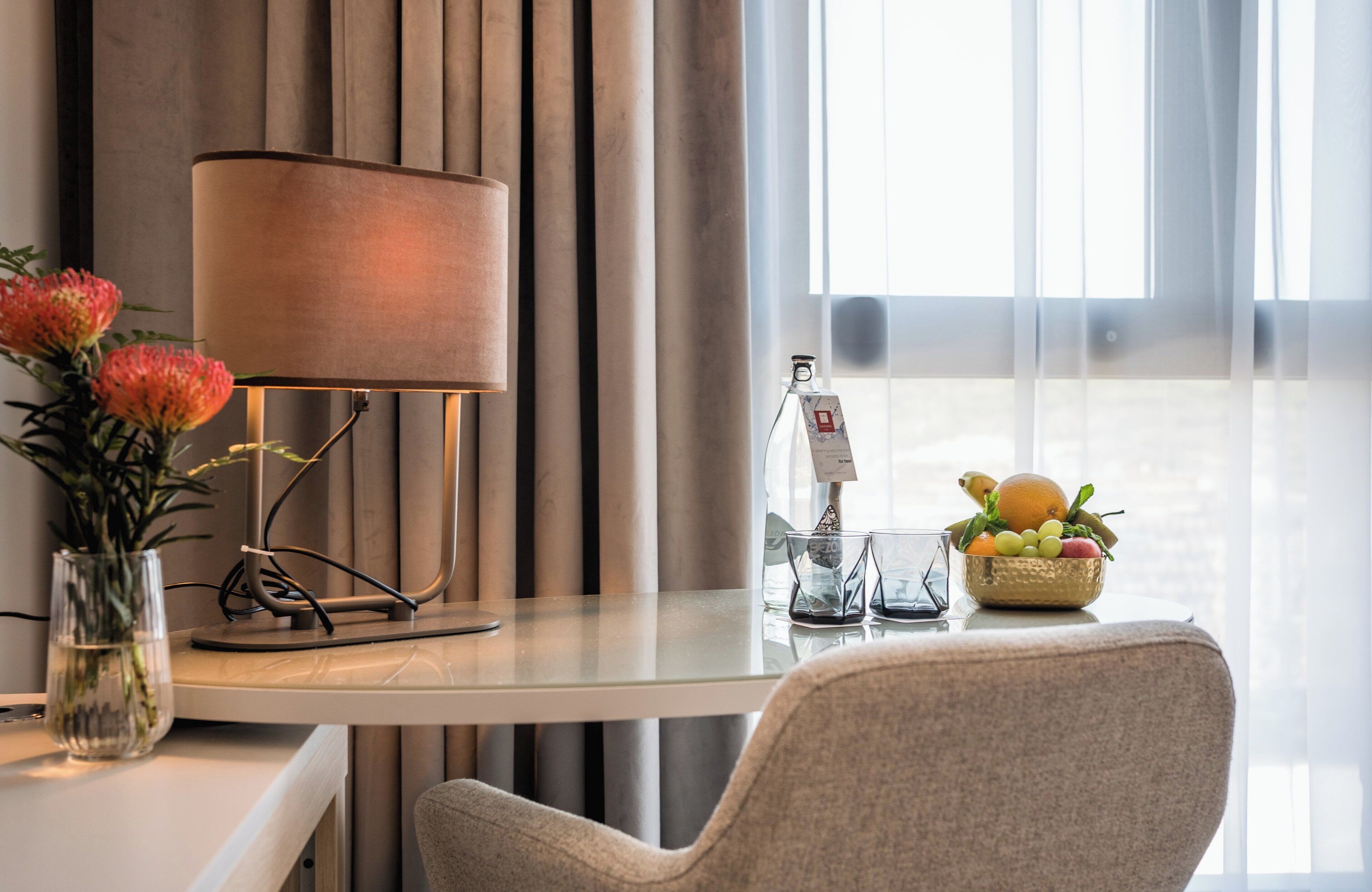 A hotel room with a desk, a lamp, a vase with flowers, a fruit bowl, and a window with curtains.