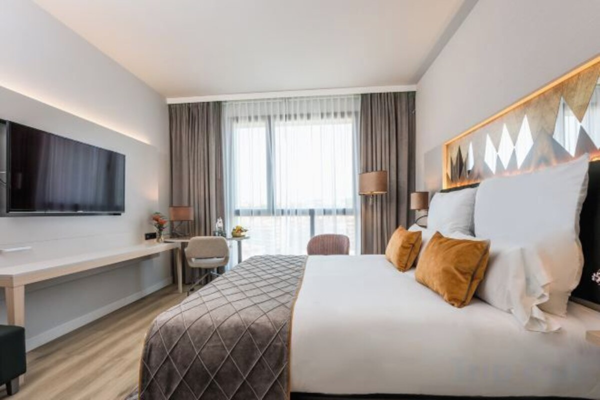 Quarto de hotel com cama, escrivaninha, televisão e janela com cortinas.