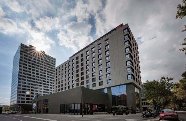 Ein modernes Hotel mit einer markanten Glasfassade, mehreren Etagen und blauem Himmel mit Sonnenschein.