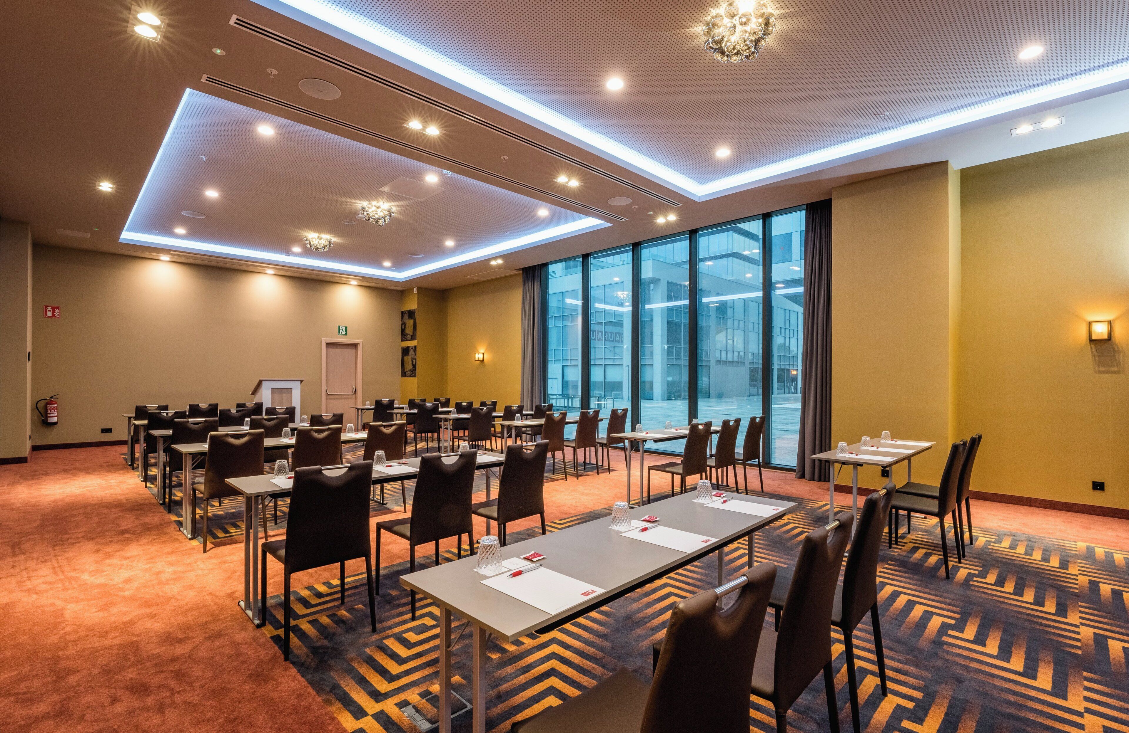 Un salón de conferencias con mesas y sillas, ventanas amplias y una alfombra estampada.