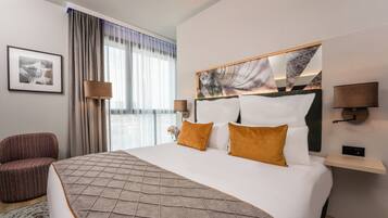 Habitación de hotel con una cama grande, cabecera decorada con obras de arte, dos lámparas de noche, una silla y una ventana con cortinas.