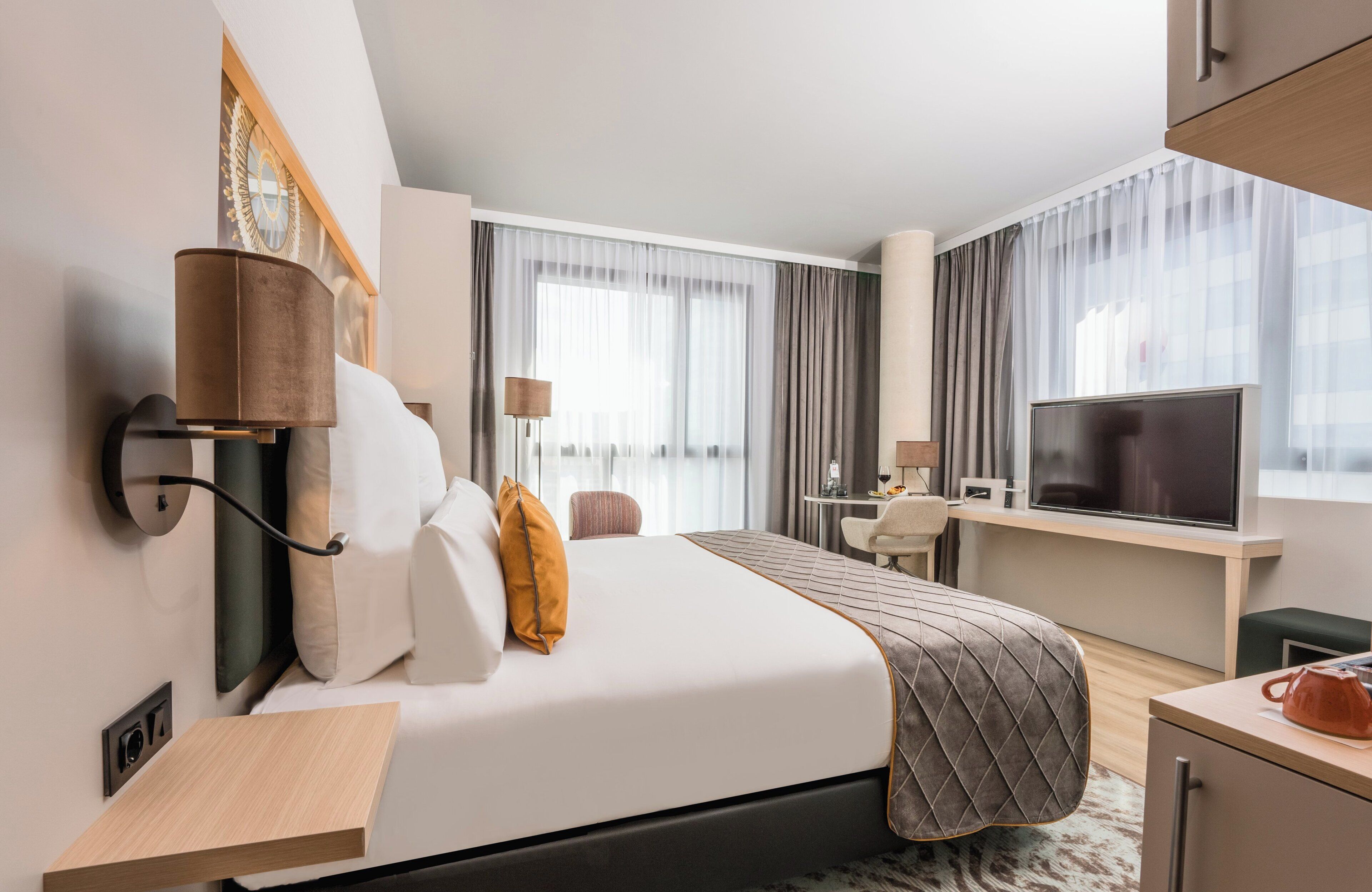 A modern hotel room with a bed, a desk, a television, and a window with curtains.