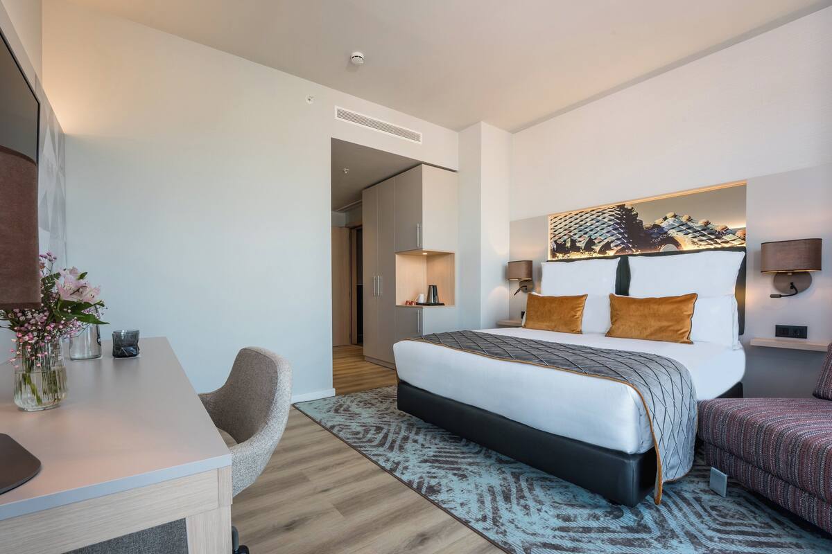 Une chambre d’hôtel moderne avec un grand lit, un bureau et une chaise. Il y a un tableau au mur et une lampe sur la table de chevet.