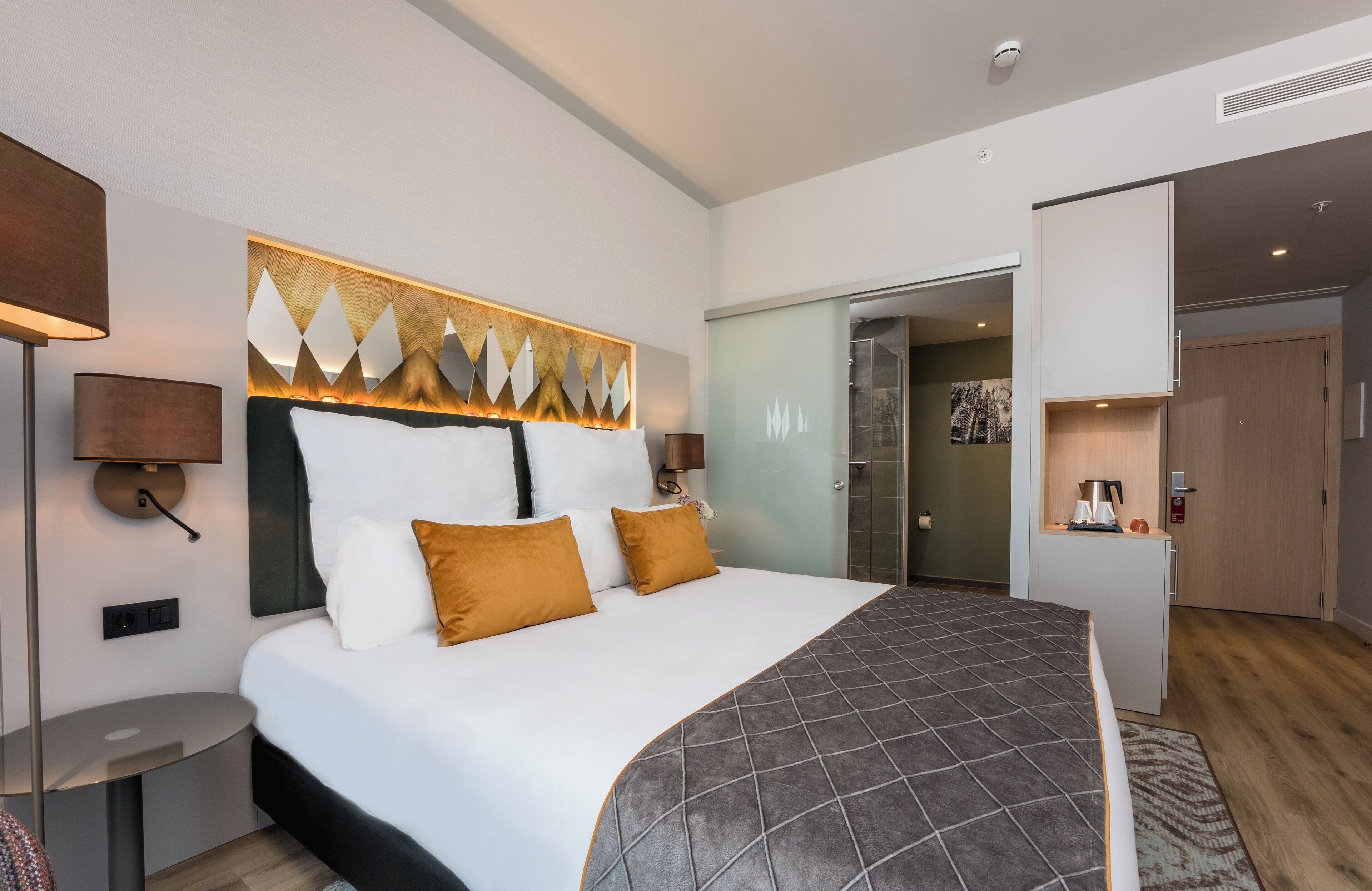 Una habitación de hotel moderna con una cama grande, lámparas de noche, una obra de arte colgada en la pared y un pequeño escritorio con lámpara.