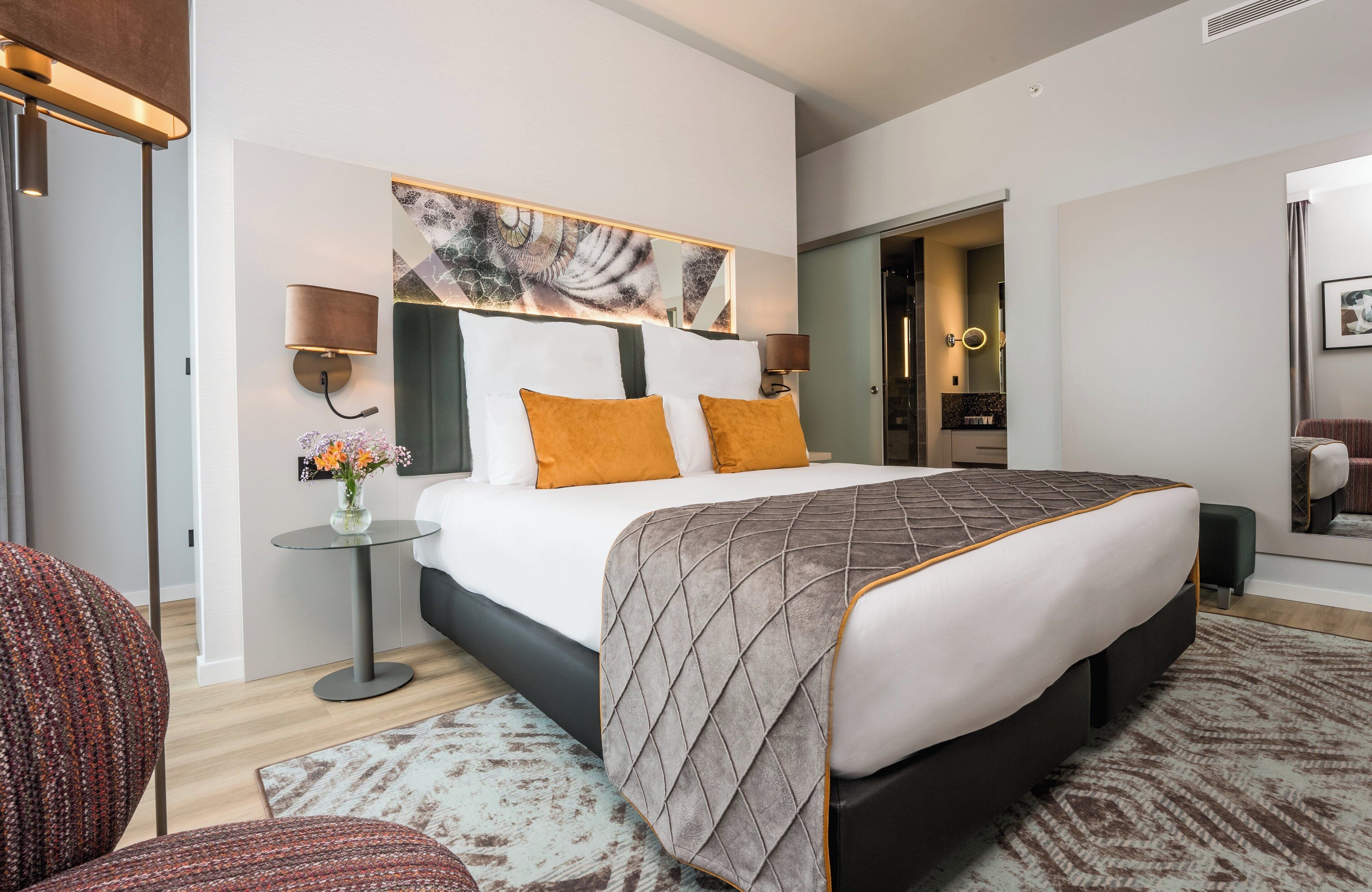 A modern hotel room with a large bed, two bedside lamps, a round side table with a vase of flowers, and a patterned rug.