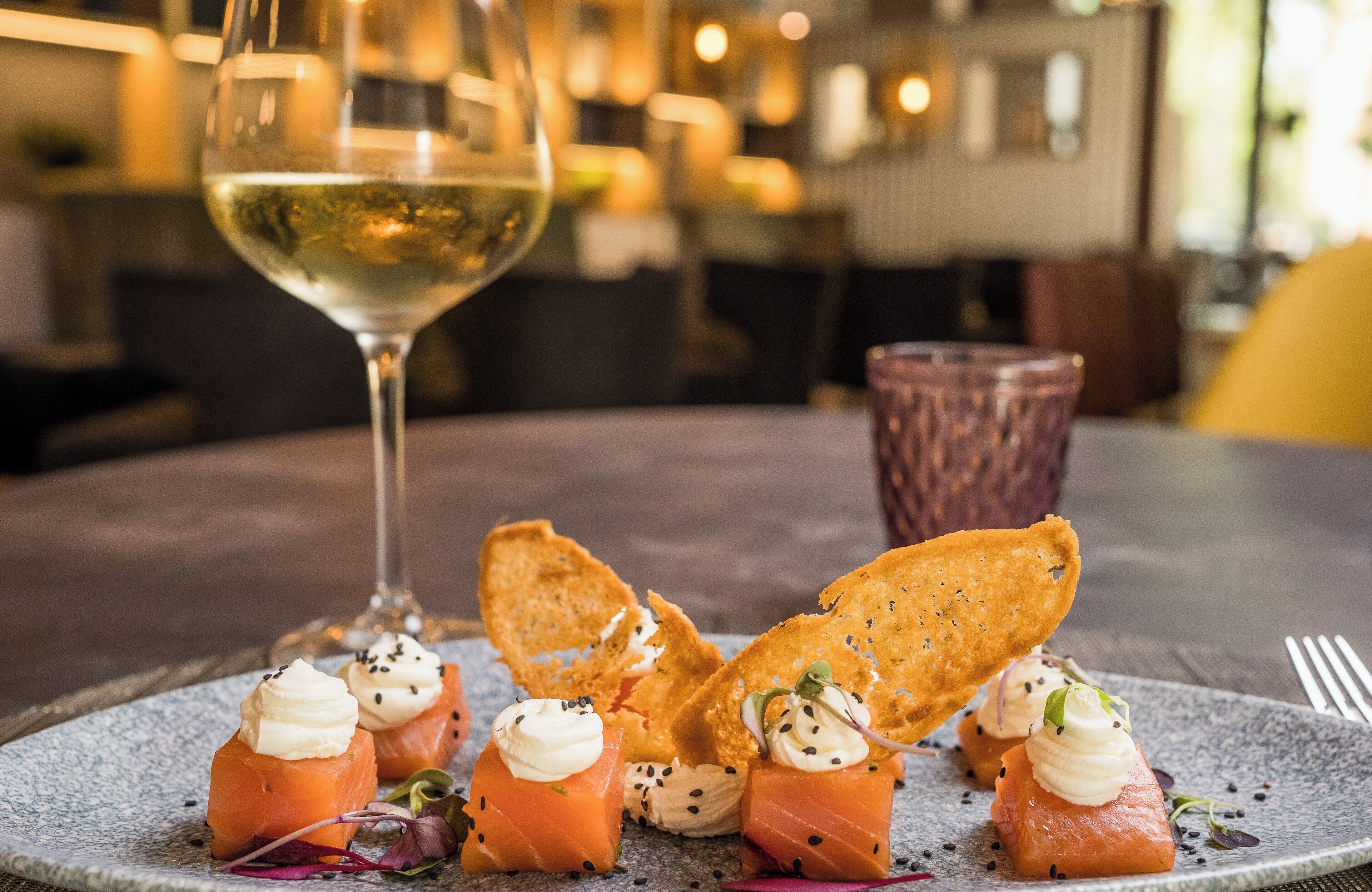 Ein Teller Vorspeisen mit Räucherlachs und Creme, dazu Cracker und Garnituren, auf einem Tisch mit einem Glas Weißwein.