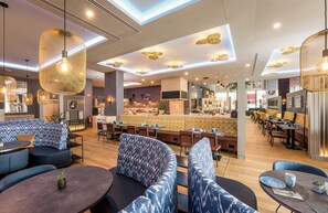Ein modernes Restaurant mit runden Tischen, blau gemusterten Sitzbänken und hängenden Pendelleuchten.