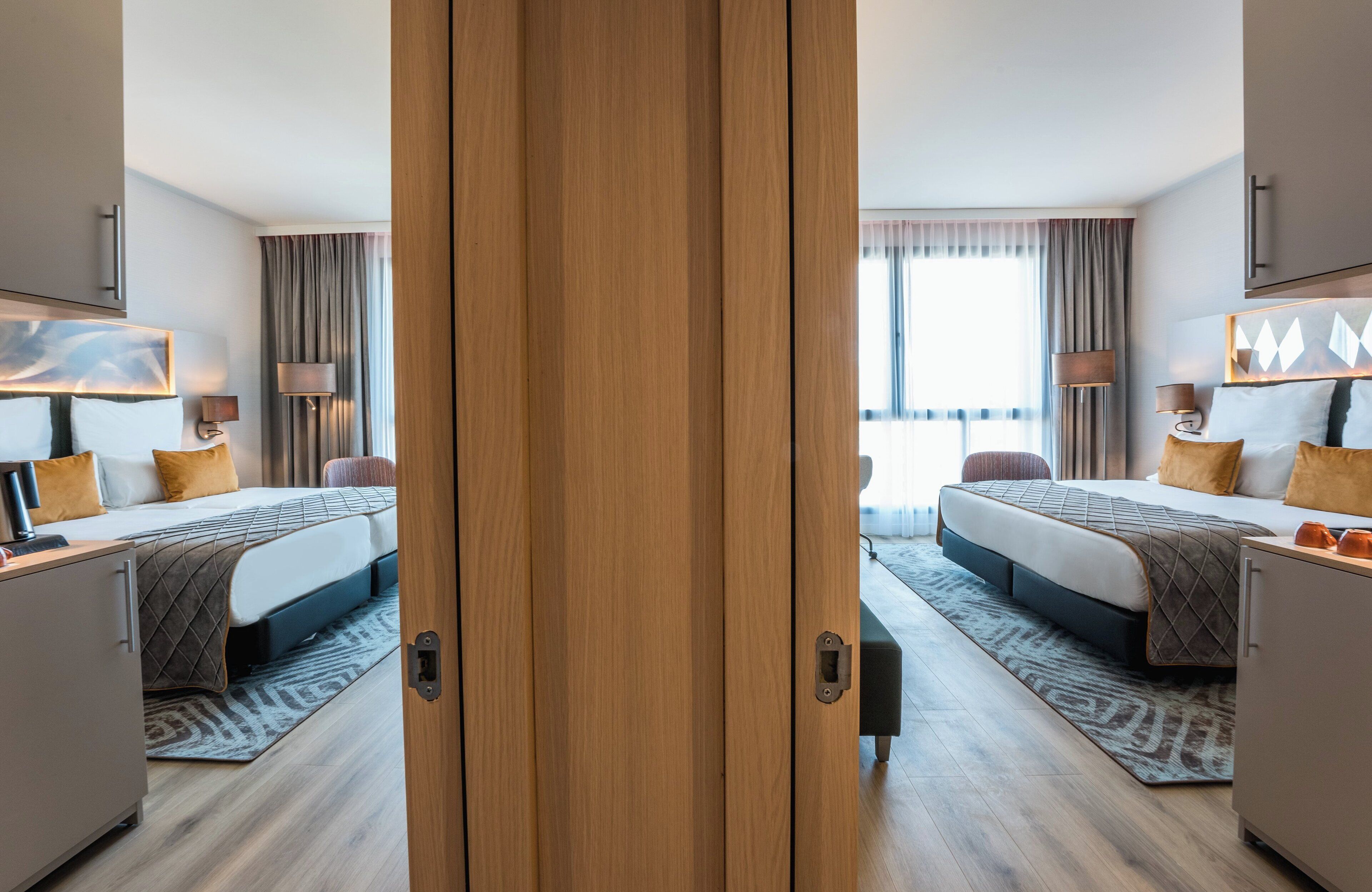 Ein Hotelzimmer mit zwei Betten, einem Kleiderschrank, einem Schreibtisch und einem großen Fenster mit Vorhängen.