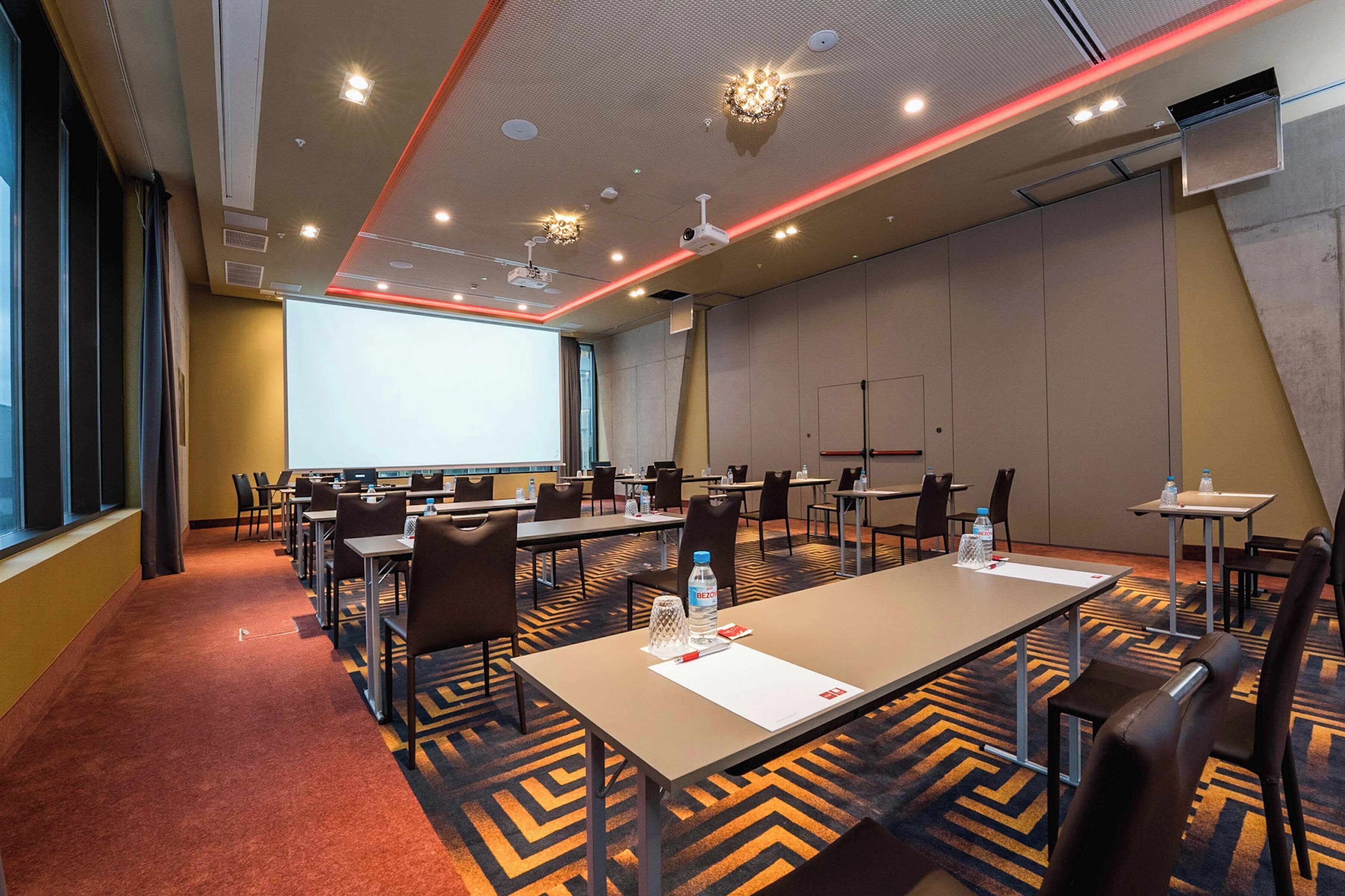 A conference room with multiple tables, chairs, a projector screen, and a large window.