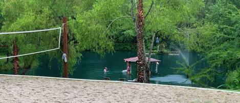 A sandy beach area with a volleyball net, a small pavilion, and a body of water surrounded by trees.