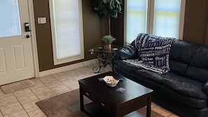 A living room with a dark leather sofa, a coffee table, a ceiling fan, and a large window with blinds. - West Wing at the Manor w/ Swimming Pond & Sand Volleyball Access (Montgomery)